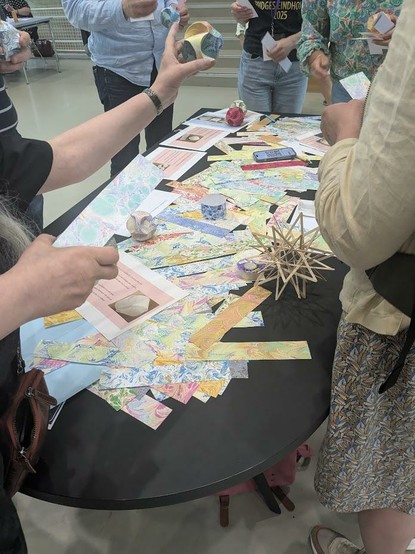 A group of people are gathered around a table filled with colorful marbled paper cut into strips and various geometric paper sculptures.