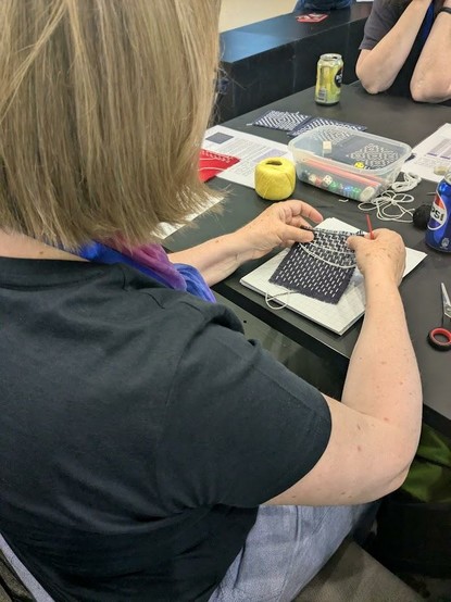 A person is focused on a craft project, seemingly weaving or embroidering a small, patterned fabric piece.