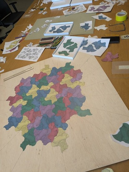 An overhead shot of a large wooden board with a complex, colorful tessellation pattern made of many interlocking, irregular shapes, alongside various art supplies.