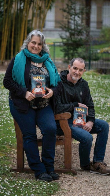 Fotografía del escritor Julio Santos y la ilustradora Patricia Pérez sentados en un banco de madera sujetando cada uno el primer libro de la colección infantil de Sara-Li: Mil años después/Mila urte geroago.
Están en un parque de la ciudad de Donostia.
El césped detrás suyo está plagado de margaritas.