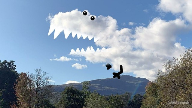 Photo predominantly of a blue sky with fluffy white clouds, with a mountain and some trees at the bottom. The clouds form a dinosaur head type shape, with wide open jaws, so I have added eyes, pointy teeth, and some little arms. It's a bit silly.